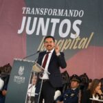 Oaxaca mayor Raymundo Chagoya speaking at a podium with "Transformando Juntos la Capital" banner behind him as people sit in antique-style carved wooden chairs watching.