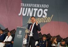 Oaxaca mayor Raymundo Chagoya speaking at a podium with "Transformando Juntos la Capital" banner behind him as people sit in antique-style carved wooden chairs watching.