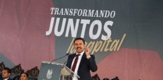 Oaxaca mayor Raymundo Chagoya speaking at a podium with "Transformando Juntos la Capital" banner behind him as people sit in antique-style carved wooden chairs watching.