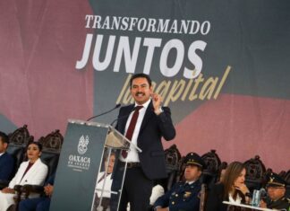 Oaxaca mayor Raymundo Chagoya speaking at a podium with "Transformando Juntos la Capital" banner behind him as people sit in antique-style carved wooden chairs watching.