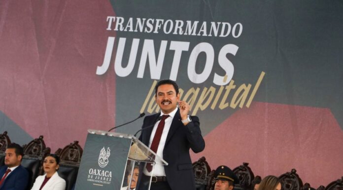 Oaxaca mayor Raymundo Chagoya speaking at a podium with "Transformando Juntos la Capital" banner behind him as people sit in antique-style carved wooden chairs watching.