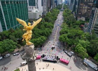 An aerial view of the Angel of Independence monument on Reforma Avenue in Mexico City, looking down the tree-lined boulevard surrounded by tall city buildings.