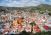 An expansive panoramic view of Guanajuato City, Mexico, showcasing its vibrant, colorful houses covering rolling hills, with prominent landmarks like the yellow Basílica Colegiata de Nuestra Señora de Guanajuato and other historic buildings, under a partly cloudy sky.
