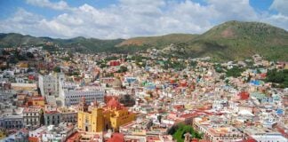 An expansive panoramic view of Guanajuato City, Mexico, showcasing its vibrant, colorful houses covering rolling hills, with prominent landmarks like the yellow Basílica Colegiata de Nuestra Señora de Guanajuato and other historic buildings, under a partly cloudy sky.