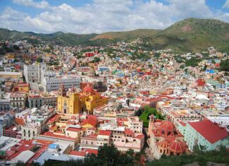 An expansive panoramic view of Guanajuato City, Mexico, showcasing its vibrant, colorful houses covering rolling hills, with prominent landmarks like the yellow Basílica Colegiata de Nuestra Señora de Guanajuato and other historic buildings, under a partly cloudy sky.
