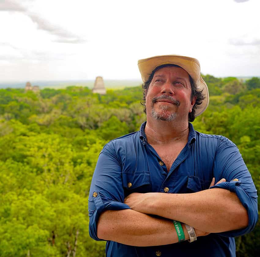 Ed Barnhart wears a wide-brimmed hat and work shirt with forest and Mexican pyramids in the background