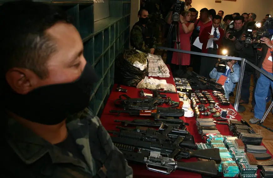 Mexican soldier in 2009 in Saltillo, Mexico standing guard over a display of Mexican pesos in plastic bags, several automatic military-grade rifles, boxes of bullets, ammunition cartridges, and jewelry confiscated from the Gulf Cartel. Reporters with cameras are crowded around the display taking photos and video footage of the the display.