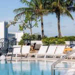 Image of a luxury hotel in a tropical location, with a rooftop pool with white deck chairs around it, palm trees behind it and buildings in the far left background with wide balconies on each floor, like a hotel.