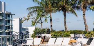 Image of a luxury hotel in a tropical location, with a rooftop pool with white deck chairs around it, palm trees behind it and buildings in the far left background with wide balconies on each floor, like a hotel.