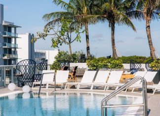 Image of a luxury hotel in a tropical location, with a rooftop pool with white deck chairs around it, palm trees behind it and buildings in the far left background with wide balconies on each floor, like a hotel.