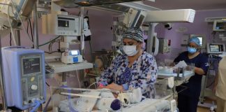 A masked doctor attends to a tiny infant in a hospital incubator