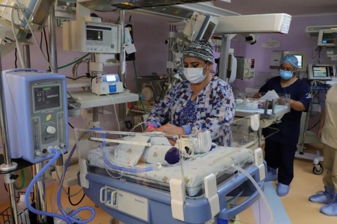 A masked doctor attends to a tiny infant in a hospital incubator