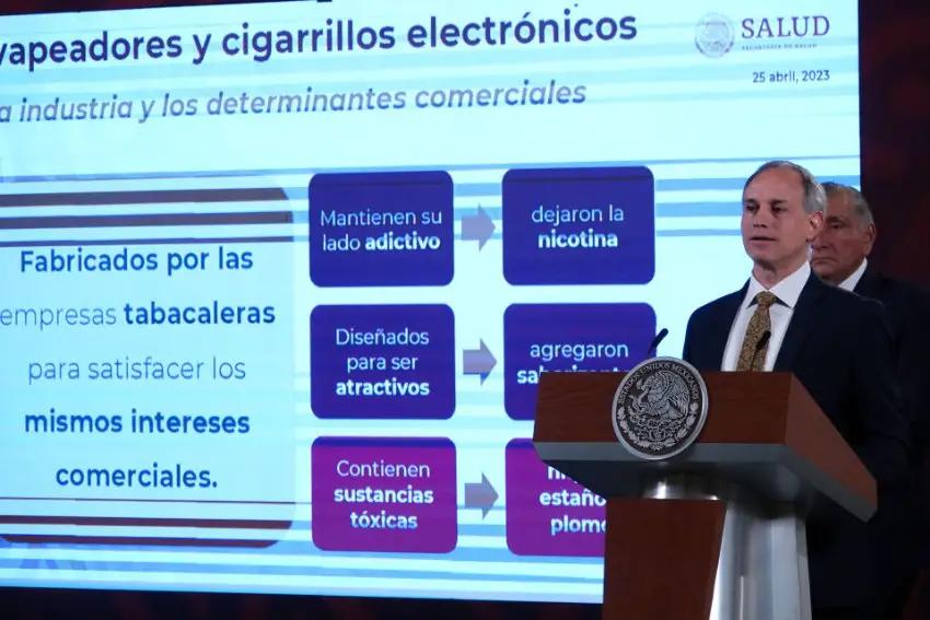 Mexico's former deputy health minister Hugo Lopez-Gatell standing at the presidential podium at a 2023 press conference in front of a projection screen with an infographic showing how the dangers of vaping and e-cigarettes are equal to that of conventional tobacco cigarettes to explain why Mexico was restricting the sale of the former items.