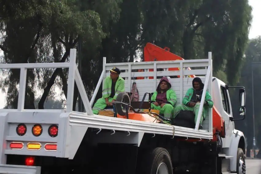 Municipal workers in bright green hazmat like jumpsuits sitting in a truck bed as they are driven on a Mexico City street on an official holiday