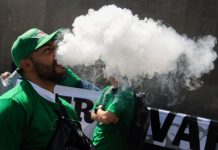 A Mexican man in a green jacket and green baseball cap stands outside the Chamber of Deputies in Mexico City, exhaling a a thick white cloud of smoking after inhaling from a vaping device. He is part of a protest to keep vaping legal.