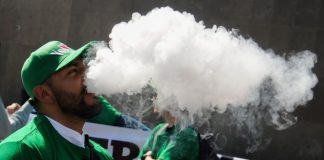 A Mexican man in a green jacket and green baseball cap stands outside the Chamber of Deputies in Mexico City, exhaling a a thick white cloud of smoking after inhaling from a vaping device. He is part of a protest to keep vaping legal.