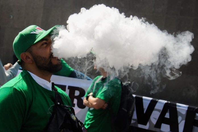 A Mexican man in a green jacket and green baseball cap stands outside the Chamber of Deputies in Mexico City, exhaling a a thick white cloud of smoking after inhaling from a vaping device. He is part of a protest to keep vaping legal.