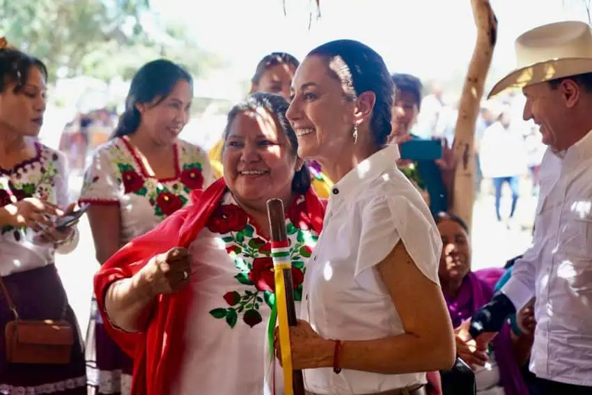 Mexico's President Sheinbaum standing with a indigenous Yaqui woman in Sonora. She is holding a traditional Indigenous scepter in her hand decorated with ribbons in green, white and yellow.