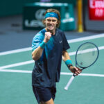 A male tennis player, Andrey Rublev, with a blue headband and wristbands clenches his fist and shouts in triumph on the court, holding his tennis racket in his other hand.