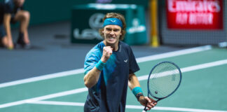 A male tennis player, Andrey Rublev, with a blue headband and wristbands clenches his fist and shouts in triumph on the court, holding his tennis racket in his other hand.