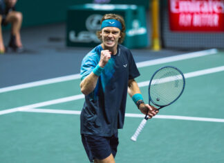 A male tennis player, Andrey Rublev, with a blue headband and wristbands clenches his fist and shouts in triumph on the court, holding his tennis racket in his other hand.