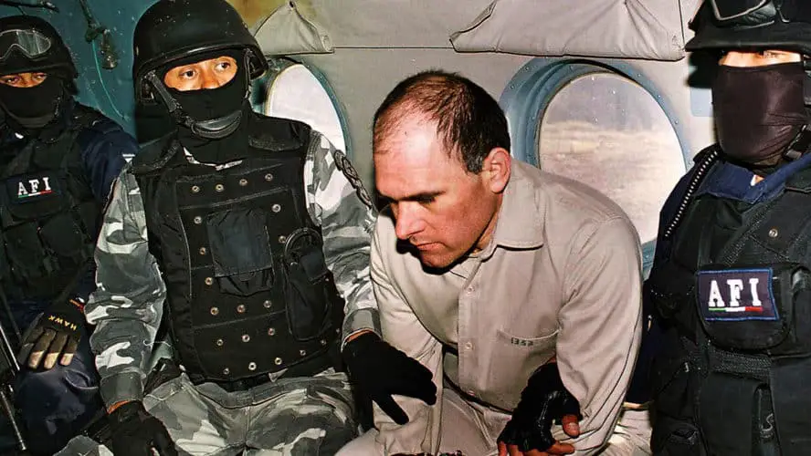 A middle aged Mexican man in a beige prison at the center of the picture is sitting under restraint inside a military plane by three soldiers in uniforms, helmets and baclavas who sit around him.