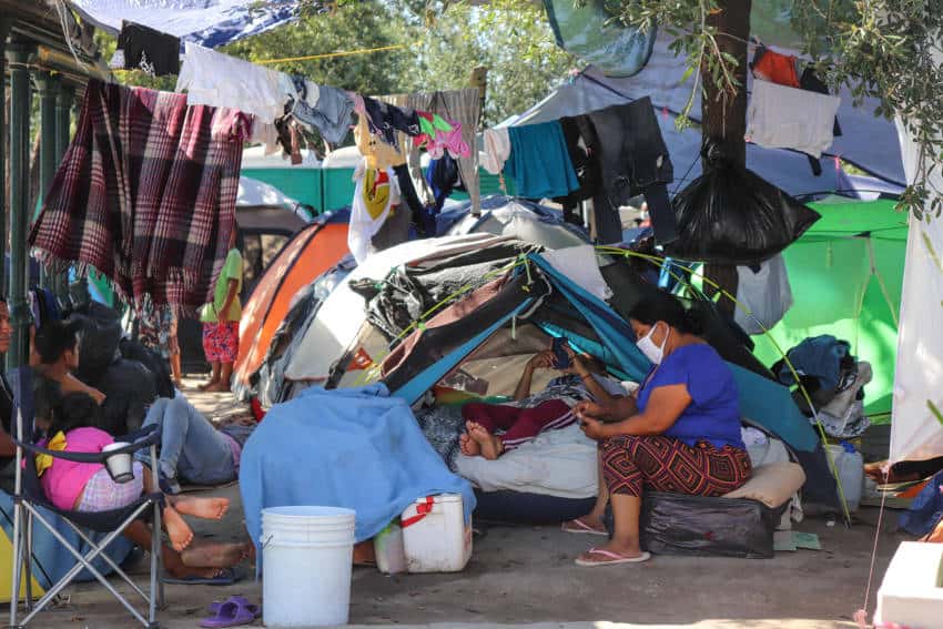 Deported migrants sitting near and sleeping in tents lined up in a row. Around them are clothes lines hanging from posts and people sitting outside in folding chairs