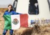 Astronaut Katya Echazarreta displays a Mexican flag in front of a Blue Origin spacecraft in a field, while wearing a blue space suit