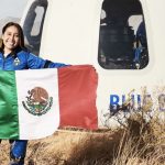 Astronaut Katya Echazarreta displays a Mexican flag in front of a Blue Origin spacecraft in a field, while wearing a blue space suit