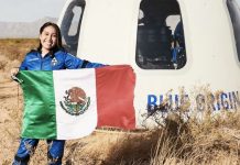 Astronaut Katya Echazarreta displays a Mexican flag in front of a Blue Origin spacecraft in a field, while wearing a blue space suit