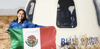 Astronaut Katya Echazarreta displays a Mexican flag in front of a Blue Origin spacecraft in a field, while wearing a blue space suit