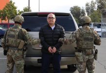 Former Gulf Cartel leader Osiel Cárdenas Guillén in handcuffs standing in front of the back of a silver SUV. He's facing the camera while two ICE employees in military fatigues are standing with their backs to the camera on either side of Cardenas Guillen. Cardenas is in a parka and black pants. He wears black framed glasses and is mostly bald.