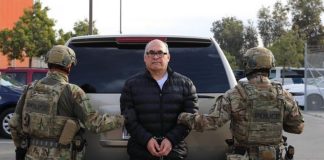 Former Gulf Cartel leader Osiel Cárdenas Guillén in handcuffs standing in front of the back of a silver SUV. He's facing the camera while two ICE employees in military fatigues are standing with their backs to the camera on either side of Cardenas Guillen. Cardenas is in a parka and black pants. He wears black framed glasses and is mostly bald.