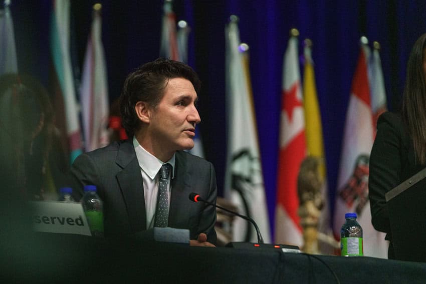 Canada's Prime Minister Justin Trudeau sitting at a panel table at a negotiation meeting. Behind him are several large flags on stands.