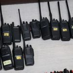 Black two way communication radios with labels on each bearing a different number written in marker, lying on a table on display.