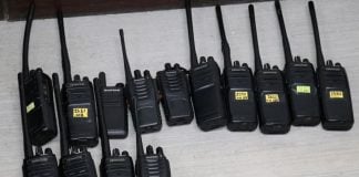 Black two way communication radios with labels on each bearing a different number written in marker, lying on a table on display.