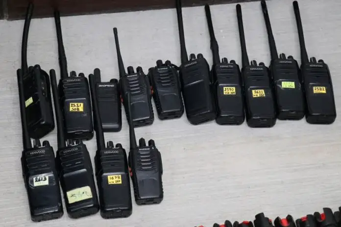 Black two way communication radios with labels on each bearing a different number written in marker, lying on a table on display.