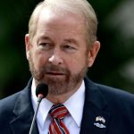 Ronald D. Johnson standing in front of a microphone at a Department of State event. On the lapel of his suit is a pin bearing the flags of the U.S. and El Salvador