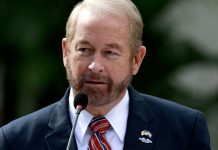 Ronald D. Johnson standing in front of a microphone at a Department of State event. On the lapel of his suit is a pin bearing the flags of the U.S. and El Salvador