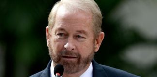 Ronald D. Johnson standing in front of a microphone at a Department of State event. On the lapel of his suit is a pin bearing the flags of the U.S. and El Salvador