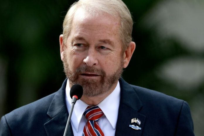 Ronald D. Johnson standing in front of a microphone at a Department of State event. On the lapel of his suit is a pin bearing the flags of the U.S. and El Salvador