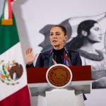 Claudia Sheinbaum standing at the presidential podium at the National Palace at her daily press conference. Her left hand is raised with her palm facing reporters as she speaks.
