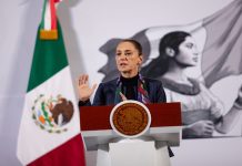 Claudia Sheinbaum standing at the presidential podium at the National Palace at her daily press conference. Her left hand is raised with her palm facing reporters as she speaks.