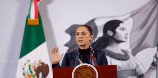 Claudia Sheinbaum standing at the presidential podium at the National Palace at her daily press conference. Her left hand is raised with her palm facing reporters as she speaks.