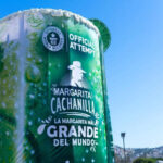 A massive 'Margarita Cachanilla' statue in Tijuana, Mexico. resembling an oversized cocktail cup, with the words "Guinness World Records Official Attempt" visible, promoting "La Margarita Más Grande del Mundo' (The World's Largest Margarita). The structure stands under a clear blue sky with the Tijuana skyline in the background.