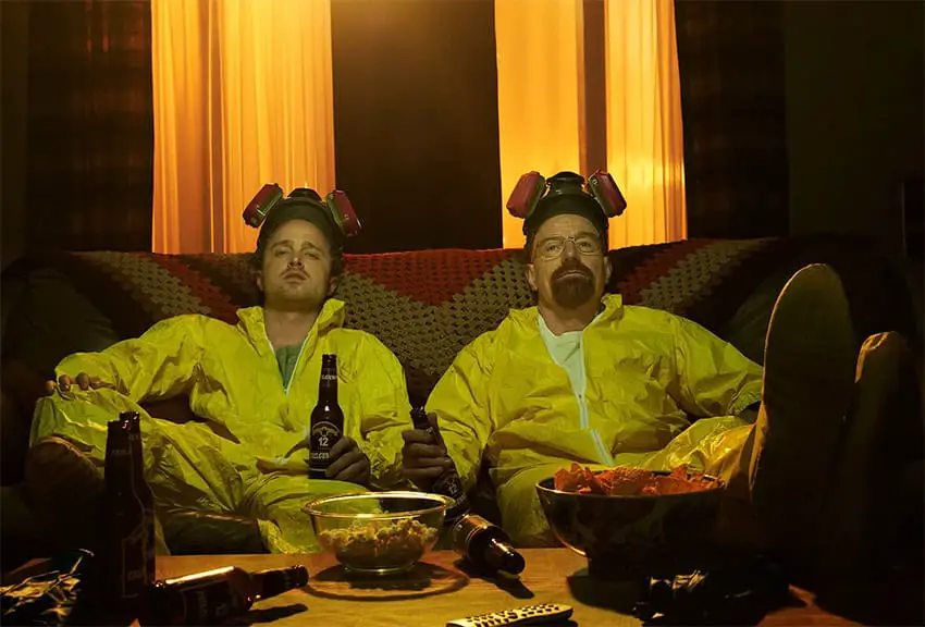 Two men in yellow chemical suits and respirators sit on a couch in front of beers and chips in a scene from the TV series Breaking Bad