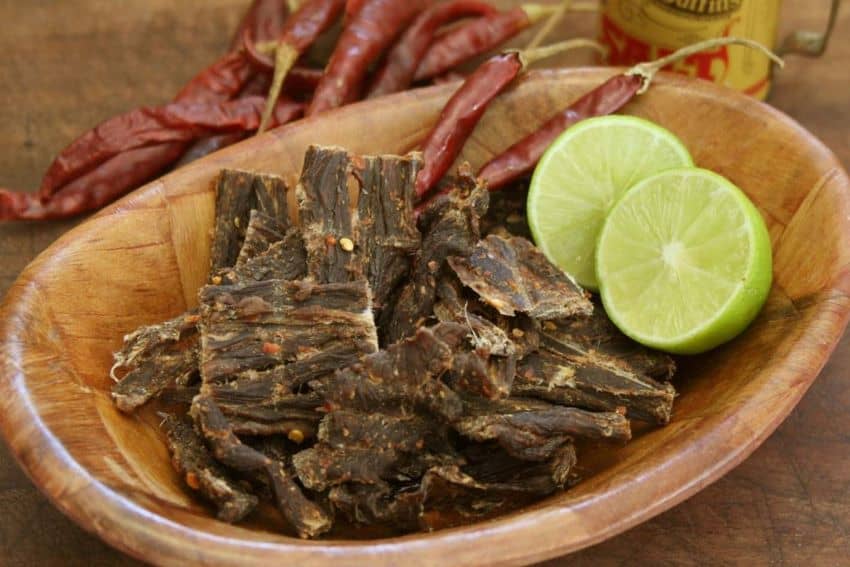 Pieces of carne seca in a bowl with red chilis and a lime split in two.