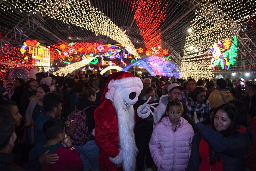 A crowd, including someone dressed as Santa with a calavera mask, inside a tunnel of Christmas lights