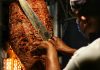 A taquero cuts meat of a trombo of al pastor taco meat.