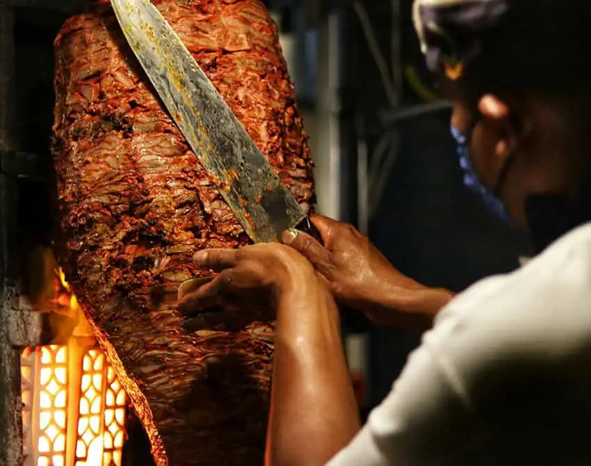 A taquero cuts meat of a trombo of al pastor taco meat.